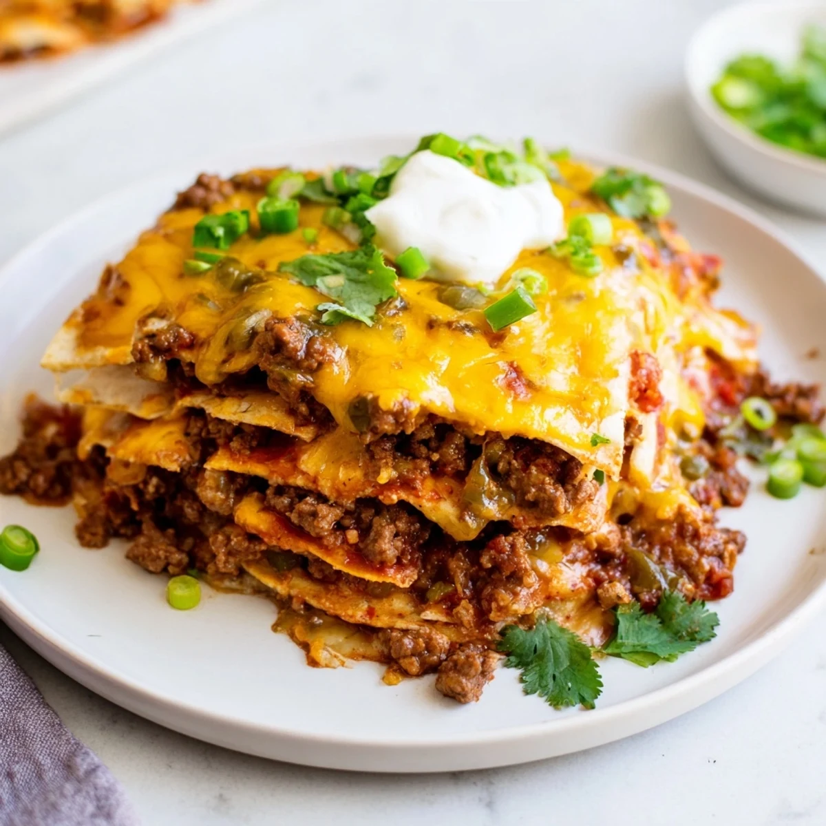 A close-up slice of Beef Enchilada Casserole reveals hearty filling with black beans and zesty red enchilada sauce, served on a white plate with a dollop of sour cream.