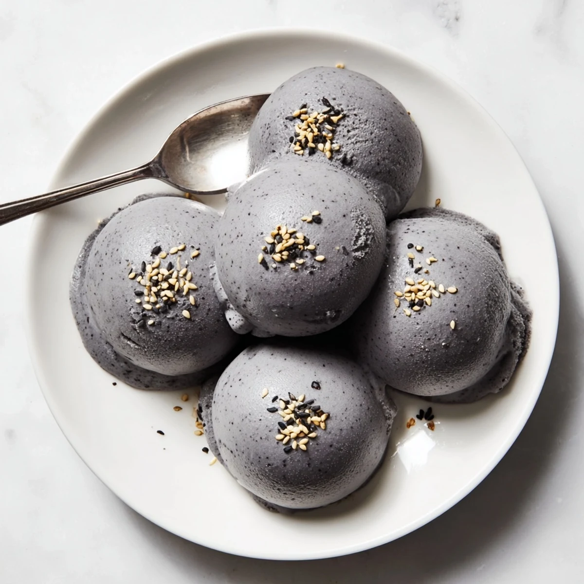 A scoop of homemade Toasted Black Sesame Ice Cream sits in a white bowl, its creamy gray hue speckled with sesame seeds and drizzled with honey.