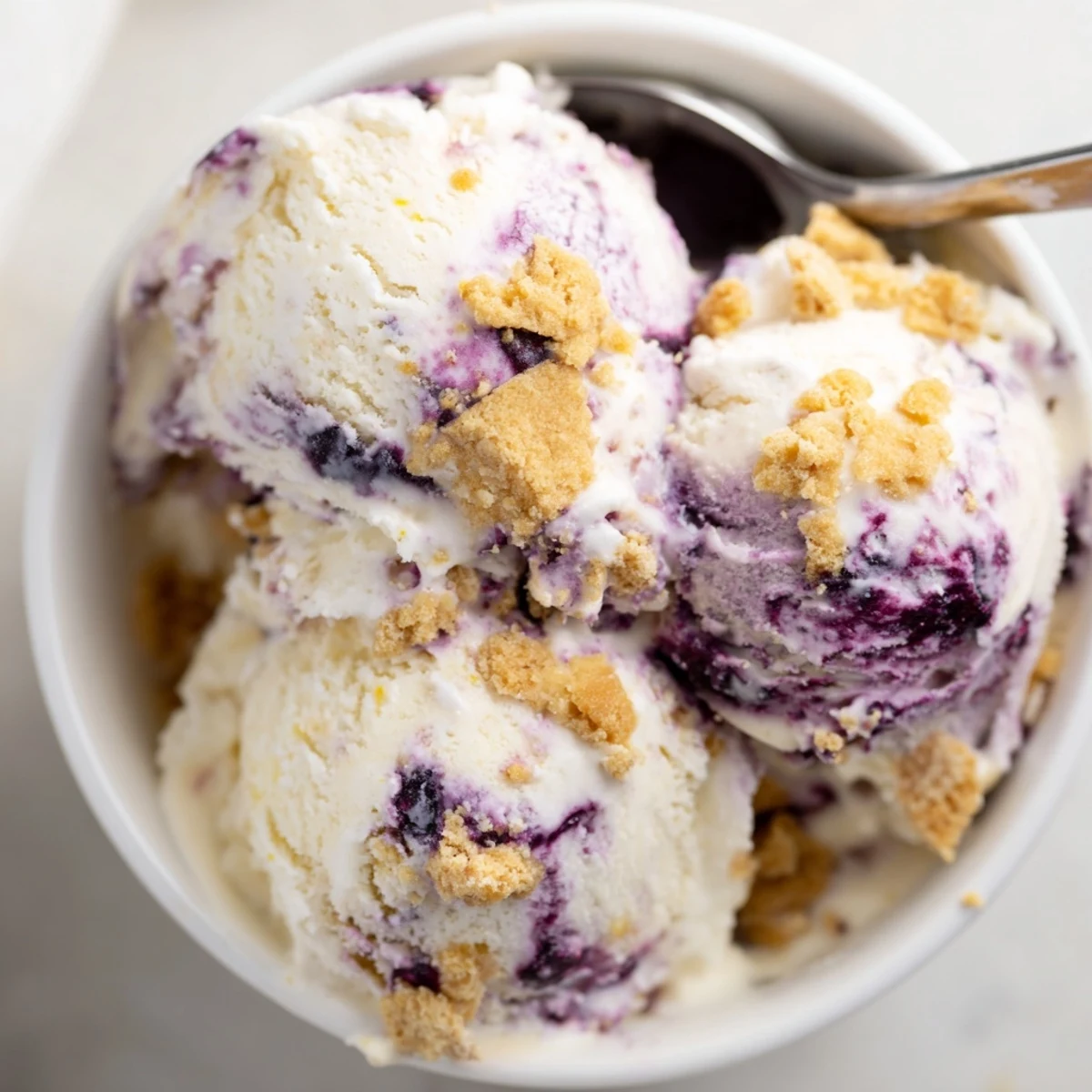 Creamy Lemon Blueberry Cheesecake Ice Cream scoops into a bowl with graham cracker crumbs and fresh blueberries.